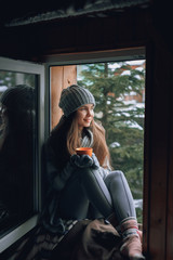 Beautiful  woman holding and drinking a cup of coffee or cocoa in gloves sitting home by the window. Blurred winter snow tree background. Morning, coziness and people concept