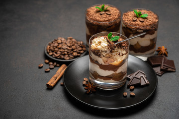 Classic tiramisu dessert in a glass cup and pieces of chocolate on stone cutting board on dark concrete background