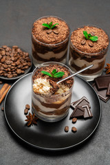 Classic tiramisu dessert in a glass cup and pieces of chocolate on stone cutting board on dark concrete background