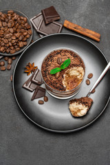 Classic tiramisu dessert in a glass cup and pieces of chocolate on dark concrete background