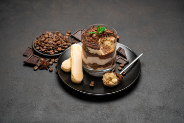 Classic tiramisu dessert in a glass cup and pieces of chocolate on dark concrete background