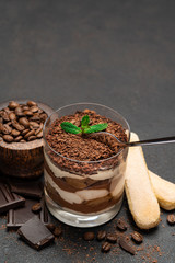 Classic tiramisu dessert in a glass cup and pieces of chocolate on dark concrete background
