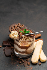 Classic tiramisu dessert in a glass cup and pieces of chocolate on dark concrete background