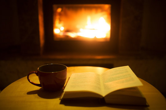 A book with a cup of hot drink on the table against the background of a burning fireplace. Cozy winter atmosphere