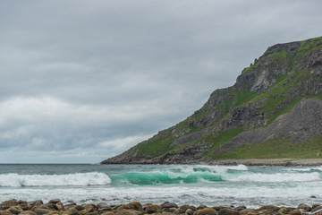 Waves at the beach. In the foreground stones and in the background a mountain. The sky is grey and cloudy and the water has a turquoise color. Unstadt Beach, Lofoten Islands, Norway.