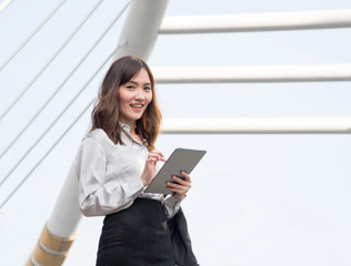 Lifestyle of a beautiful young Asian businesswoman holding digital tablet while standing in the city. Happy working girl smile and  using internet outdoor over building background.