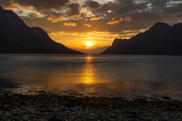 Fototapeta premium Golden sunset at Norwegian fjord with mountains. Sun shines on clouds above and reflects in the water. Dark mountain silhouttes. In foreground stone shore. Ersfjordbotn, Tromso, Norway.