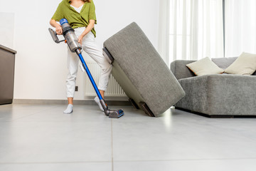 Young woman cleaning floor under the sofa with cordless vacuum cleaner at home. Concept of easy cleaning with a wireless vacuum cleaner