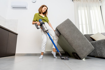 Young woman cleaning floor under the sofa with cordless vacuum cleaner at home. Concept of easy...