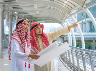 Arabian engineer talking about saudi job and verify the blueprint with commitment to success at construction site. Arab foreman in traditional clothing kandura standing on city. construction concept.