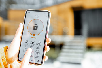 Holding phone with launched alarm app on the house background outdoors. Concept of controlling and managing home security from a mobile device