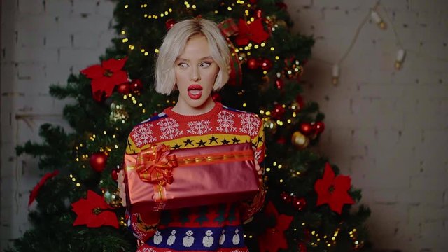 Beautiful blonde girl with short curly hair with white skin and red lipstick smiling holds in her hands bright gift boxes next to the Christmas tree indoors