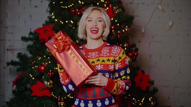 Attractive emotional young blonde woman with curly hair and red lips in a bright sweater shows gift boxes shopping next to the Christmas tree indoors