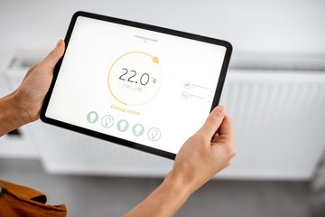 Controlling radiator heating temperature with a tablet, close-up with radiator on the background. Concept of a smart home and mobile application for managing smart devices at home - obrazy, fototapety, plakaty