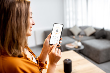 Young woman holding smart phone with launched security application at home. Concept of controlling and managing home security from a mobile device - obrazy, fototapety, plakaty