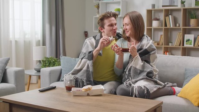 Young couple watching tv and drinking coffee with donuts hiding in a blanket