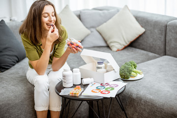 Woman taking vitamins or nutritional supplements in the form of pills while sitting on the couch at home. Concept of biohacking and preventive medicine - obrazy, fototapety, plakaty