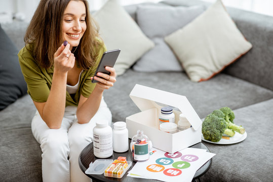 Woman Taking Nutritional Supplements In The Form Of Pills While Sitting With Smart Phone At Home. Concept Of Individual Online Selection Of Food Supplements. Preventive Medicine