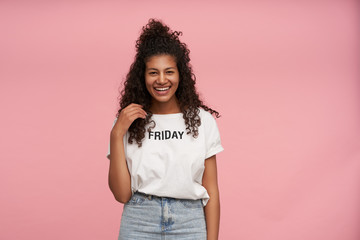 Attractive young curly long haired brunette female with dark skin touching her hair and looking positively to camera with charming smile, standing over pink background in casual clothes