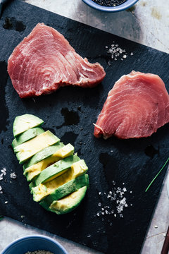 Fresh raw tuna steak ready to be prepared  for cooking on a stone lavic plate  with pink salad and avocado slice on a plate top view over a wooden cutboard background