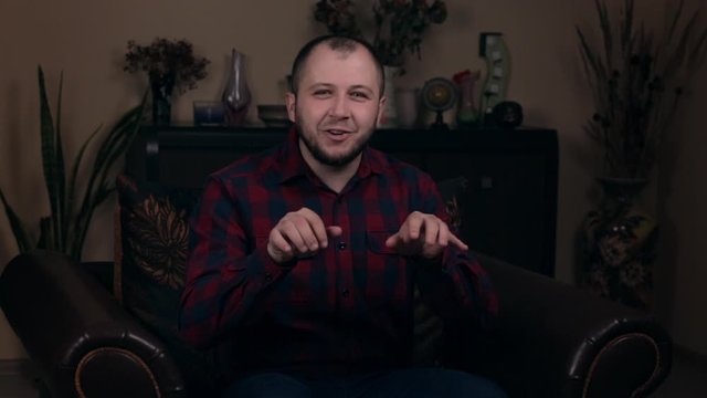 The influential Bearded Young guy, a blogger, of European appearance, sits in a chair in a plaid shirt and asks to write comments under the video. Social Media Blogging Concept.