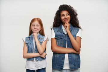 Pretty young couple of positive ladies holding raised hands on their chins and looking aside dreamily, thinking about something pleasant and smiling gently while posing over white background