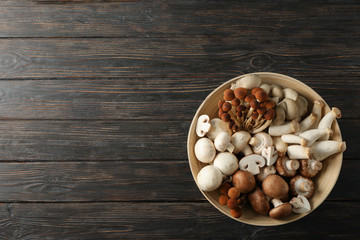 Different mushrooms in bowl on wooden background, top view
