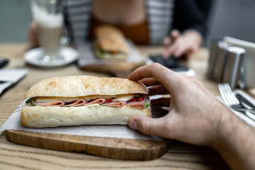 Big ham sandwich in a man's hand on the background of a girl in a restaurant. Fast food at lunch time.