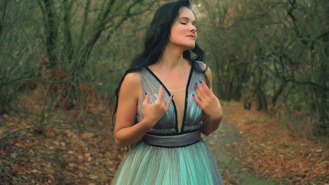 woman with long black hair in blue aqua color evening magnificent dress, gently stroking herself posing for camera. princess enjoys autumn nature. Background forest with bare trees falling leaves