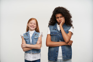 Gloomy young curly brunette female with dark skin looking sadly at camera and leaning her head on raised hand, posing over white background with cute cheerful redhead little girl