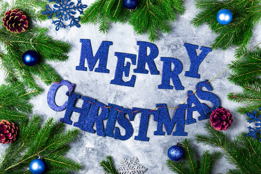 Christmas Frame made from Xmas Tree Branches, Blue Baubles, and Blue Letters Merry Christmas on the Grey Background, Top View, Flat lay