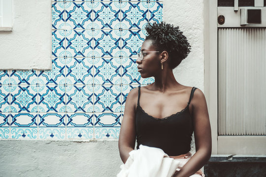 Portrait of a young charming African female in spectacles and a tank top standing next to the wall tiled with bluish traditional Portuguese Azulejo ceramic tiles and pensively looking aside