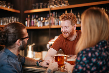 Cheerful friends leaning on bar counter, drinking beer and chatting with bartender. Night out.