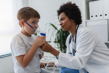 Woman African American doctor general practitioner helping child to put nebulizer inhaler face mask. Asthma treatment for children.