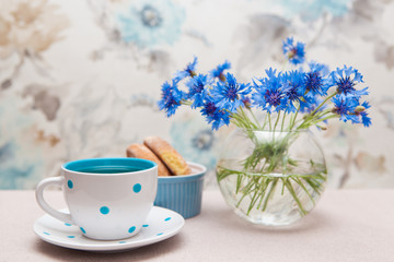 Tea in cup with dots, cookie and vase of cornflower flowers, still life