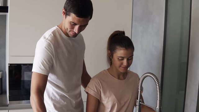 Caucasian loving couple wash fresh fruits in sink before eating in the morning on the modern kitchen. Man in jeans gently stroking the back of his caring wife then biting the red apple