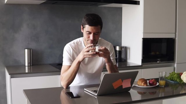 Portrait of a handsome relaxed caucasian man in a white shirt sitting on kitchen in front him laptop and smartphone. Guy sipping a black tea from transparent mug and peacefully smiling