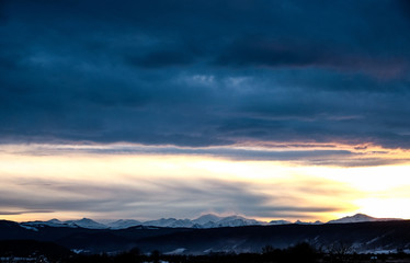 Sunset over the snow-capped mountain peaks of the Caucasus in cloudy weather in winter. The rays of the sun at sunset break through the clouds.