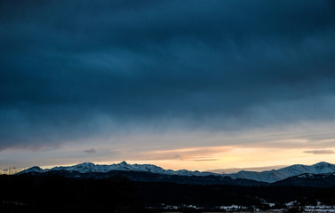 Obraz premium Sunset over the snow-capped mountain peaks of the Caucasus in cloudy weather in winter. The rays of the sun at sunset break through the clouds.