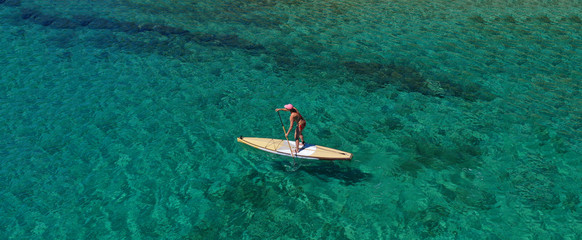 Aerial drone ultra wide photo of fit unidentified woman practising in SUP board or Stand UP Paddle surf board in tropical exotic island turquoise sea