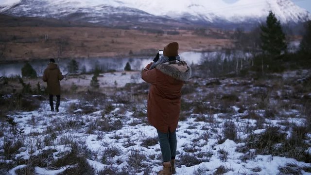 Two tourists walking in slow motion through the countryside towards the snowy mountain peaks on the background. A person on a winter coat trying to take a photo of beautiful Norway nature