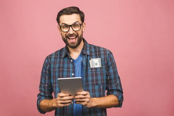 Happy winner! Young rich man in casual holding money dollar bills and tablet computer with surprise isolated over pink wall.