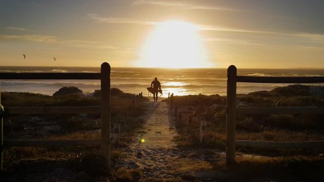 Silhouette kite surfer walks out of see on beach wooden path. Sunset behind on ocean horizon. Wind blows over big waves. Kitesurfers rides Cape Town waves. Twilight dusk evening magical time.