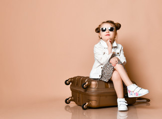 Stylish baby kid girl in leopard print dress, denim jacket and sunglasses is sitting on roller suitcase at copy space
