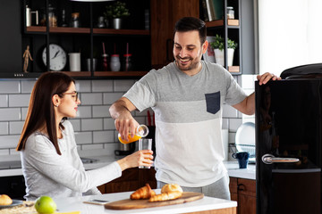 Couple in the kitchen eat breakfast with orange juice and pastry at table and he open refrigerator