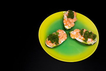 A dish of avocado halves with shrimp, sauce and herbs, a parsley leaf on a green yellow plate on dark black background. Salad of avocado and seafood, shrimp. Healthy food, fats. Omega 3 and 6.