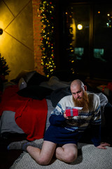 A man with a long red beard sits on the floor without pants in a winter sweater. man is holding a wrapped present on the background of New Year decorations and lights. Christmas tree. Parody, humor.