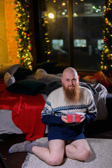 Obraz na płótnie Canvas A man with a long red beard sits on the floor without pants in a winter sweater. man is holding a wrapped present on the background of New Year decorations and lights. Christmas tree. Parody, humor.