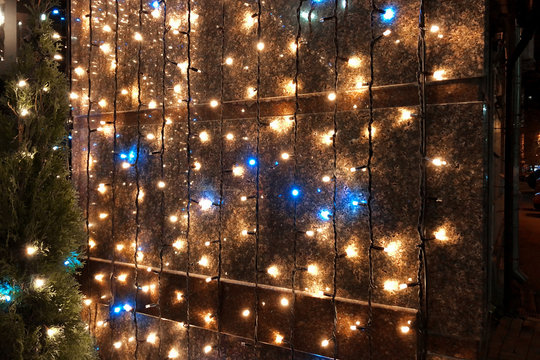 Blue And Yellow Christmas Lights Hanging On The Wall Near The House. Christmas Lights From A Garland On The Wall Before The New Year. New Year Decoration In The Evening Time.