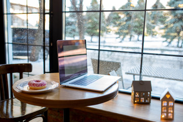 Workplace with a laptop. A table with a large window, coffee and a donut. Co-working. Workspace in a cozy coffee shop. It is snowing outside the window. Candlesticks. Work on a laptop in winter